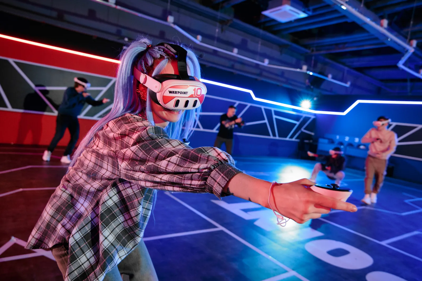 A young woman in a WARPOINT headset aims a controller in the arena while the team plays around her